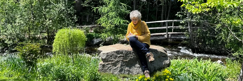 Cheryl writing beside a creek in a green garden
